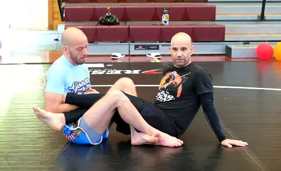 Two grapplers sitting in front of one another with their legs entangled and one players foot trapped in the other player's armpit.